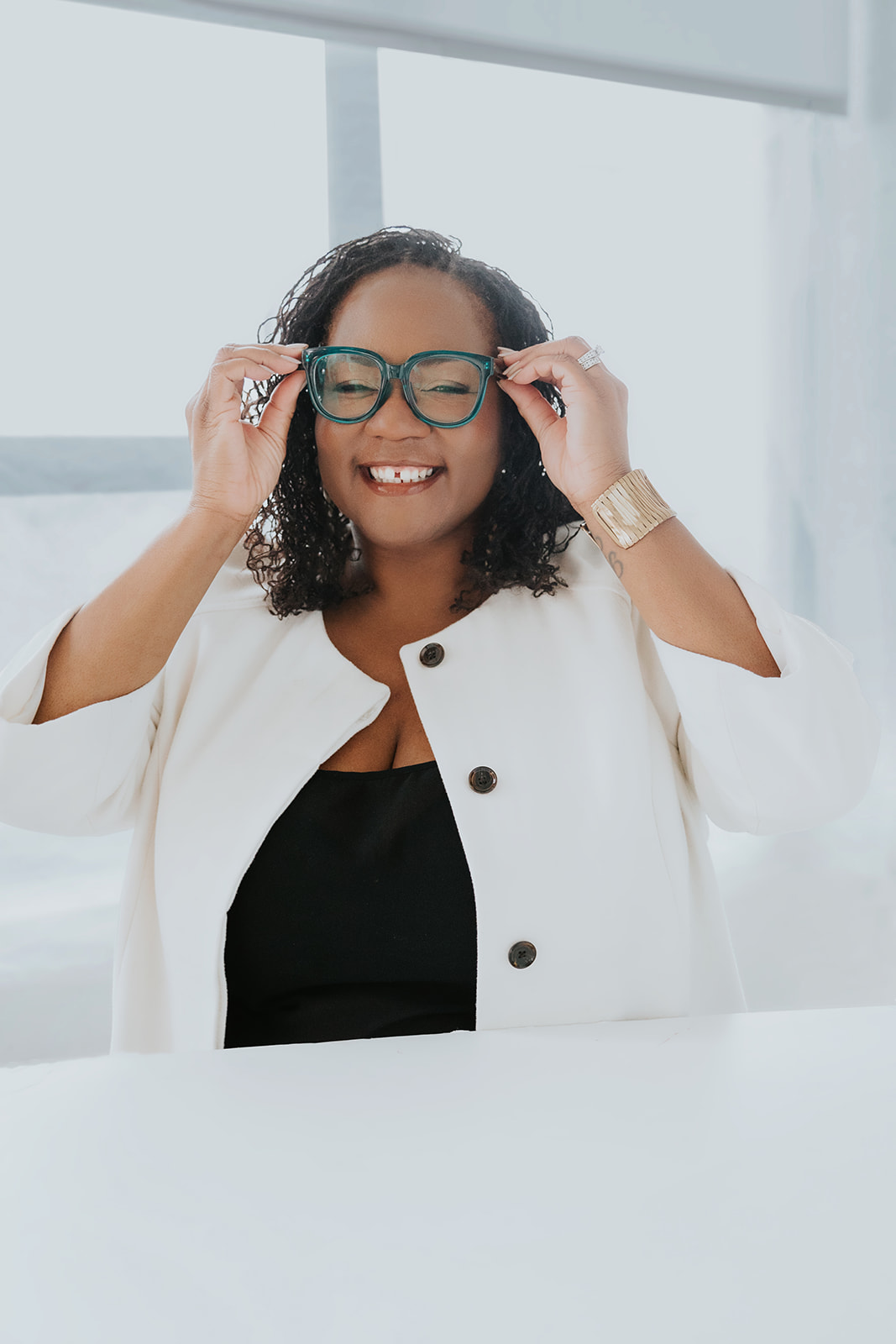 BHWEL founder Shontay Delalue, a Black woman with curly sisterlocs wearing a white jacket and a black camisole, hol her hands on either side of her teal eyeglasses as she smiles at the camera.