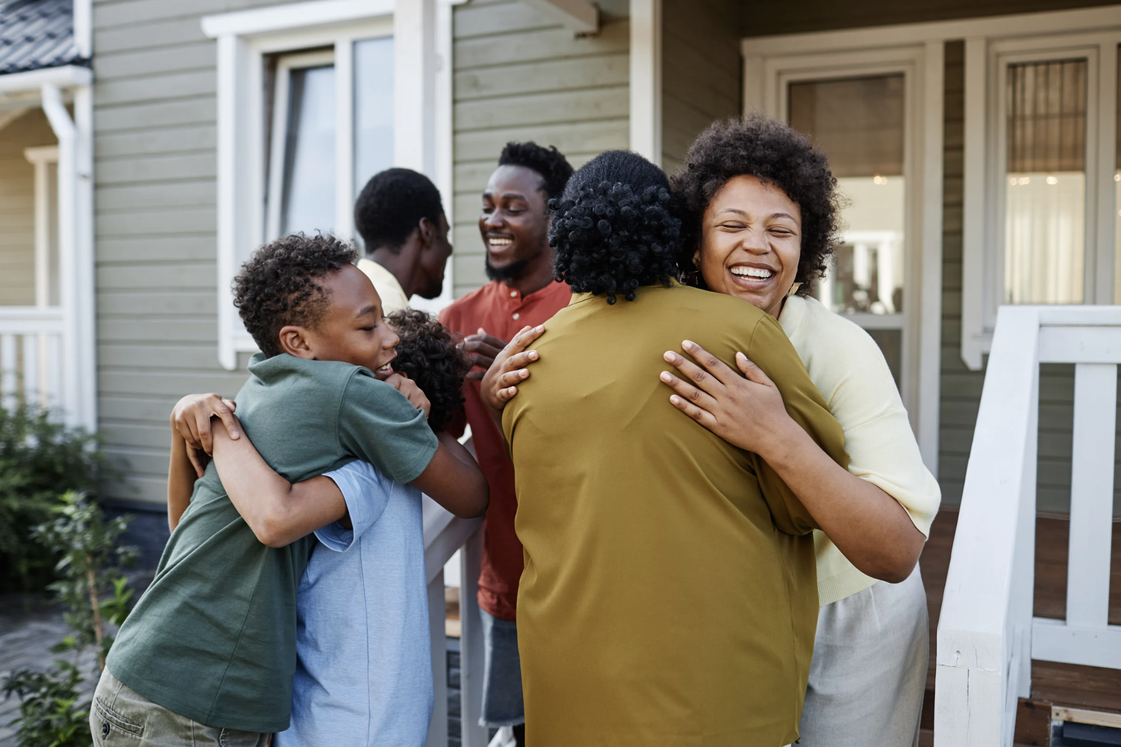 A group of six Black adults and children greeting and hugging one another just outside the front door of a home.